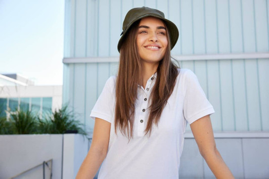 A smiling woman wearing an olive green fisherman hat and a white polo shirt outdoors, with a modern building in the background.